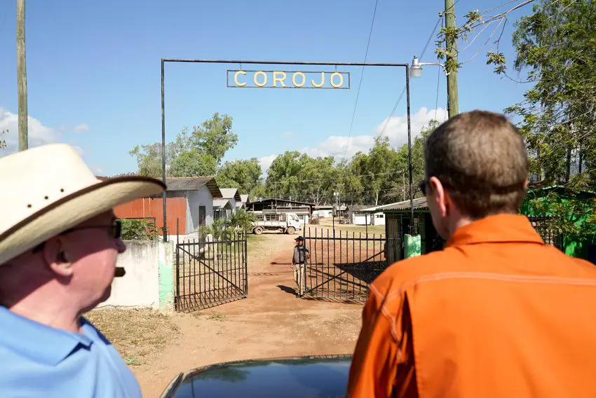 Two men standing in front of a gate with the sign 'COROJO,' leading to a tobacco farm surrounded by trees and buildings.