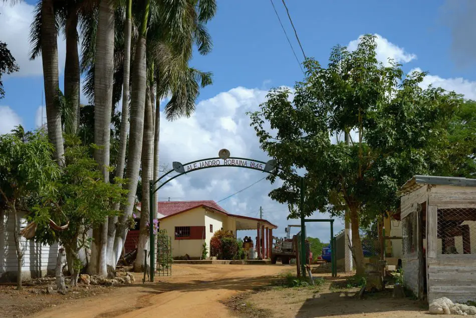 Entrance to Alejandro Robaina tobacco farm, featuring palm trees, a welcoming gate, and a rustic building under a blue sky.