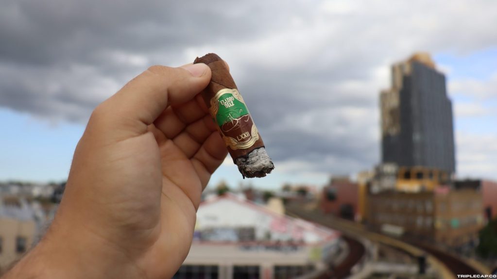 Eladio Díaz 72 Aniversario Conclusions A person holding the Eladio Díaz 72 Aniversario cigar outdoors, with a city skyline and cloudy sky in the background.