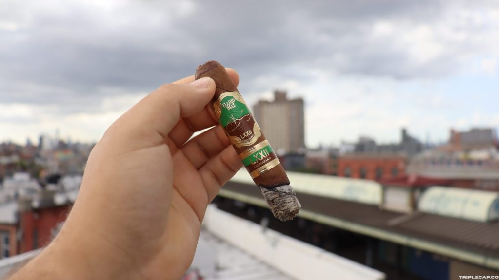 Eladio Díaz 72 Aniversario Final Third A person holding the Eladio Díaz 72 Aniversario cigar in front of a city skyline with cloudy skies.