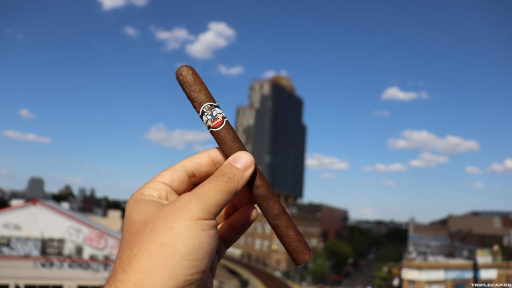 Martinez Hangry Prelight A hand holding a Martinez Hangry against a backdrop of a blue sky with scattered clouds and a city skyline.