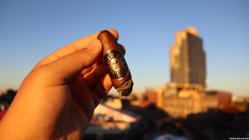 Infinity Cigars Day Shift Conclusions A hand holding a cigar with a sun design on the band, set against a backdrop of a city skyline during sunset.