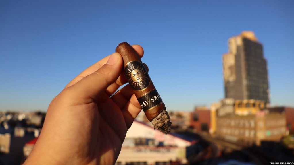 Infinity Cigars Day Shift Final Third A hand holding an Infinity Cigars Day Shift cigar against a clear blue sky with buildings in the background.