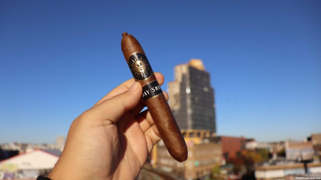 Infinity Cigars Day Shift Prelight A person holding a cigar against a clear blue sky with a city skyline in the background.