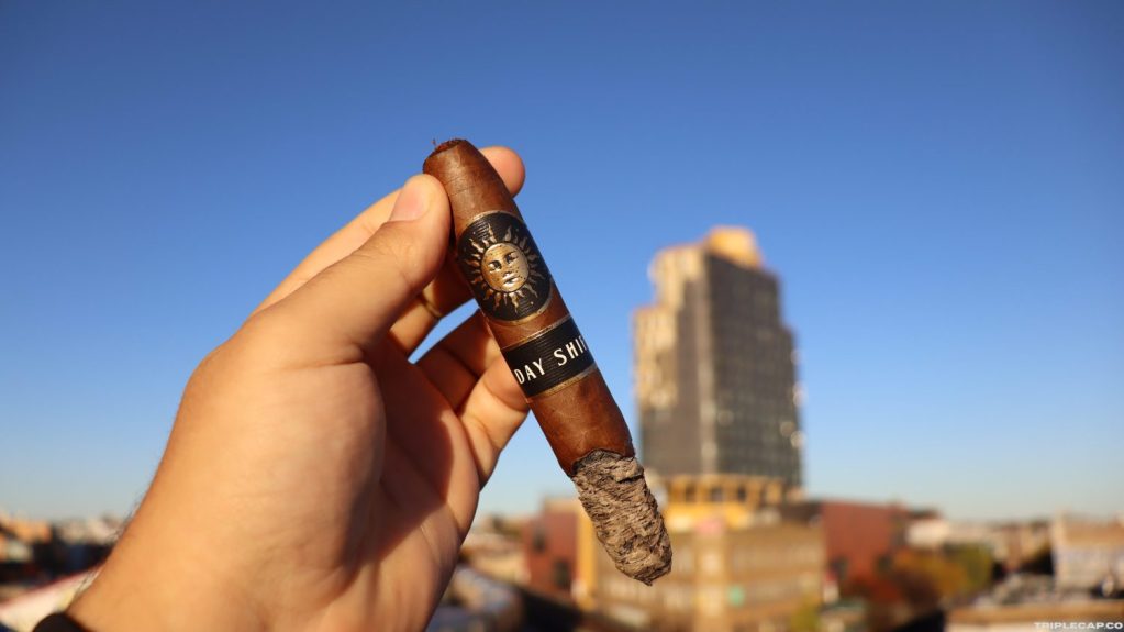 Infinity Cigars Day Shift Second Third A hand holding an Infinity Cigars Day Shift cigar against a clear blue sky and a city skyline in the background.