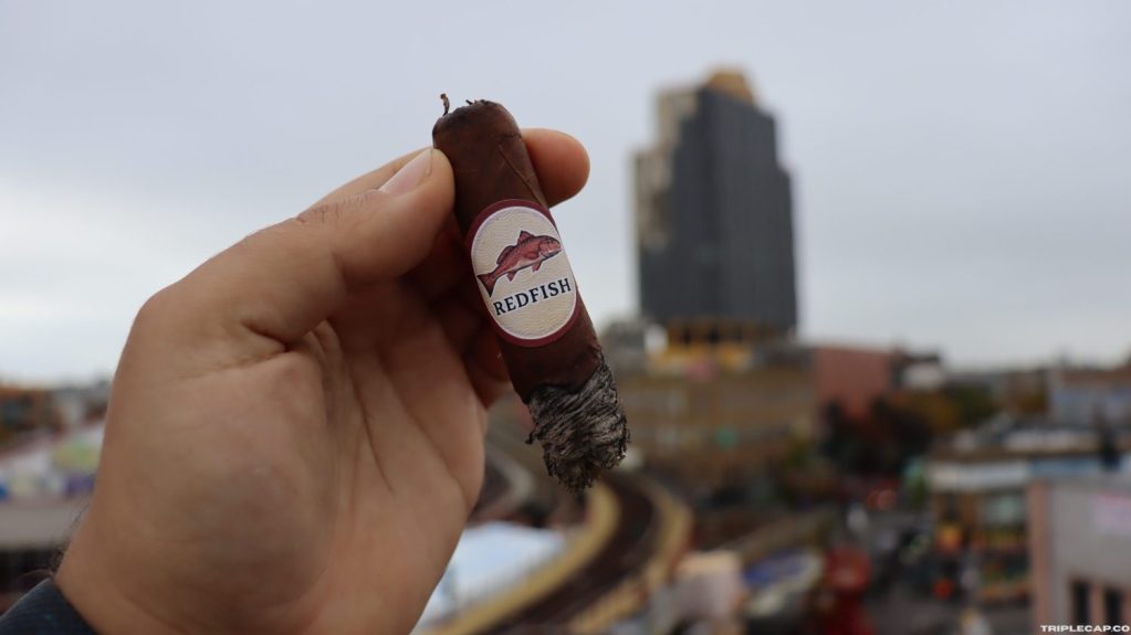 A close-up of a hand holding a cigar labeled 'Redfish' against a blurred urban background.