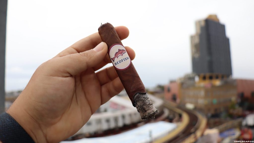 A person holding a REDFISH cigar with a cityscape in the background, showcasing a partially lit cigar and urban environment.