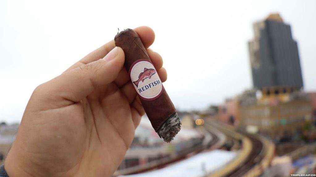A hand holding a 'Redfish' cigar against a blurred cityscape background.