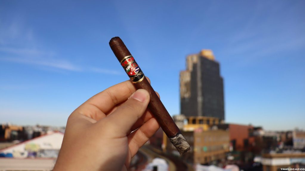 A person holding a dark brown cigar with a decorative band against a clear blue sky, with urban buildings in the background.