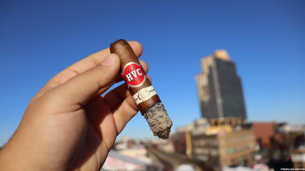 A person holding an HVC cigar in front of a city skyline with a clear blue sky.