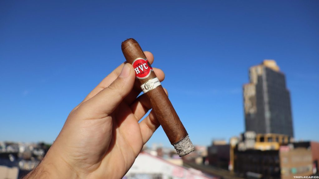 A person holding an HVC cigar against a clear blue sky, with a city skyline in the background.